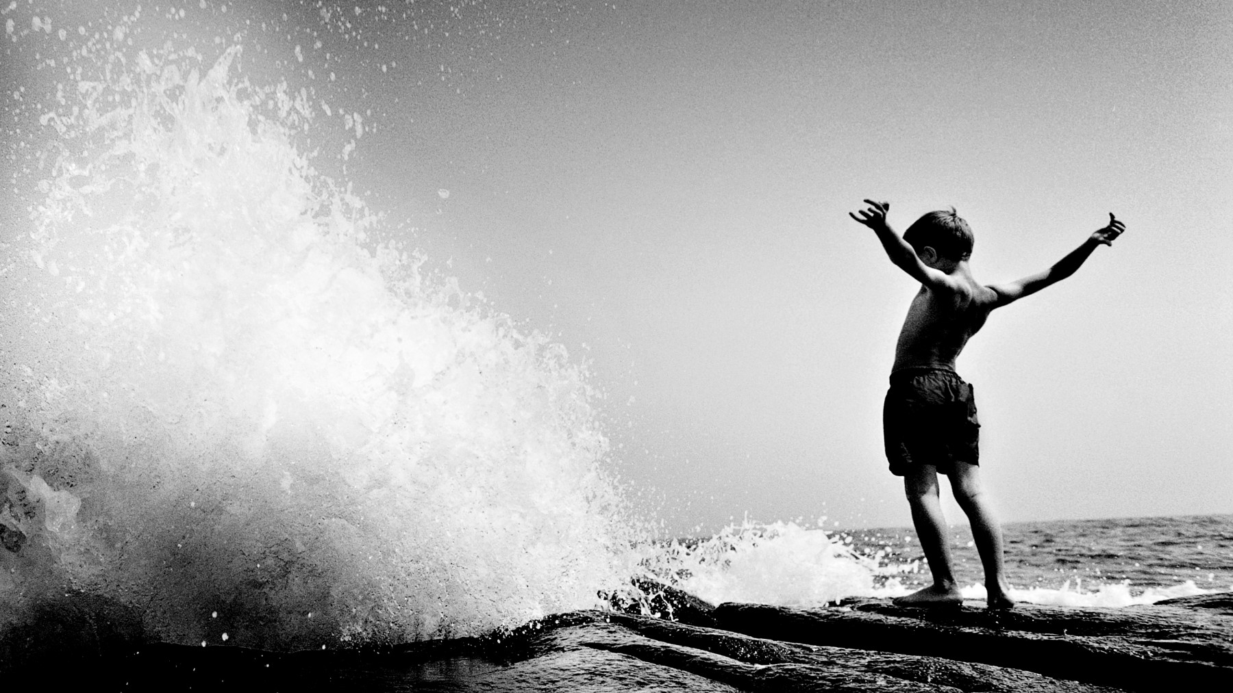 A black and white photo of a boy standing on a rock in front of a wave. Boston digital marketing agency main image. 